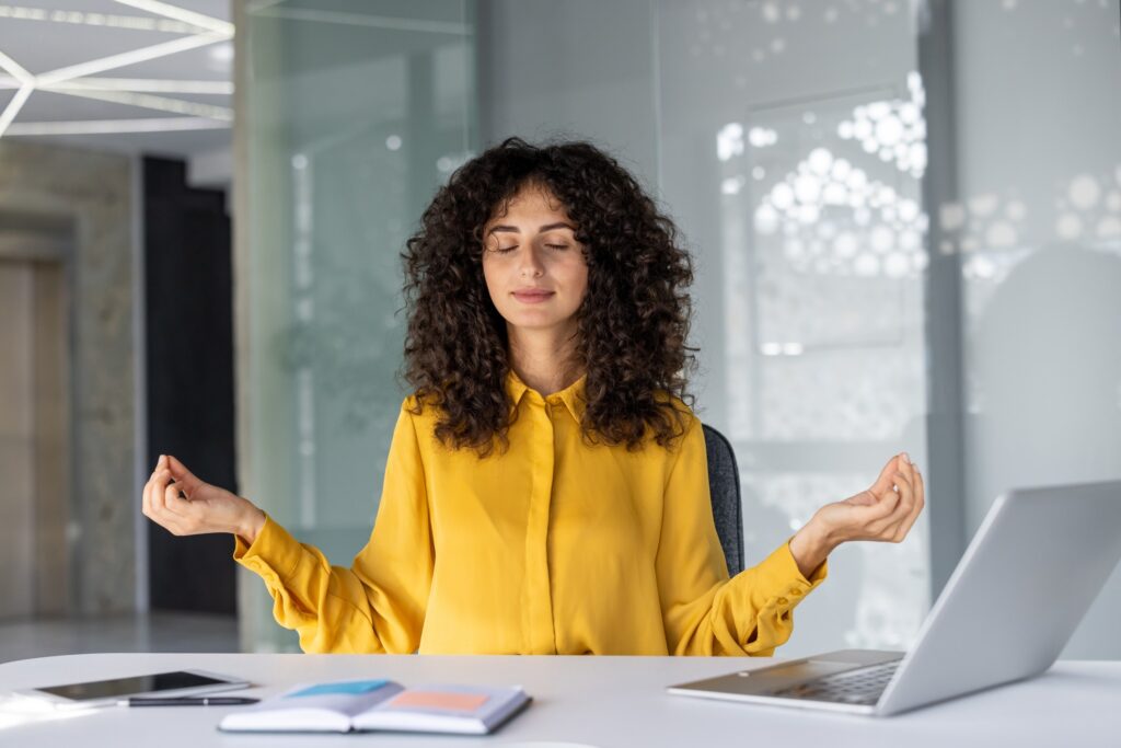 Mujer joven meditando en el escritorio de la oficina con los ojos cerrados. Practicar la atención plena y la relajación para aliviar el estrés durante el trabajo. Laptop, notebook cerca, promoviendo la calma y la concentración en un ambiente ajetreado.
